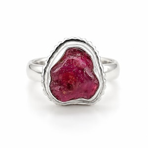 Puede incluir: Un anillo de plata con una piedra preciosa en bruto de color rojo magenta intenso, engastada en un bisel de metal texturizado y festoneado. La banda es lisa y pulida, contrastando con la superficie cristalina irregular de la piedra.