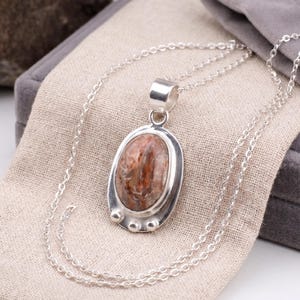 Puede incluir: Collar con colgante ovalado de plata. El colgante presenta una piedra pulida de forma ovalada con tonos marrones y crema, engastada en un bisel de plata. El collar se exhibe sobre una tela beige.