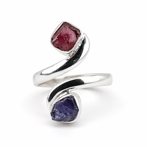 Op de afbeelding: Een ring in bypass-stijl gemaakt van gepolijst zilver, met twee open uiteinden. Het ene uiteinde bevat een ruwe, dieprode edelsteen, terwijl het andere uiteinde een onbewerkte, donkerblauwe edelsteen vasthoudt. De metalen band buigt soepel rondom.