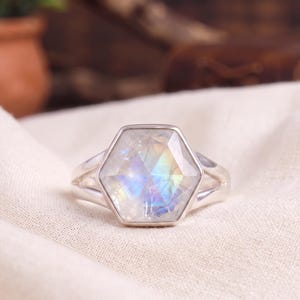 May include: A silver ring featuring a large, faceted, hexagonal-shaped moonstone with iridescent blue and rainbow flashes. The gemstone is set in a polished silver bezel, mounted on a split-shank silver band that tapers toward the back.