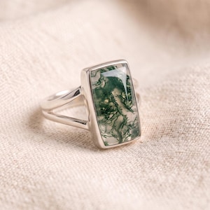 Könnte beinhalten: Ein Silberring mit einem rechteckigen Moosachat-Edelstein, der grüne, farnartige Einschlüsse aufweist und in einer glatten Metallfassung sitzt. Die Ringschiene hat ein geteiltes Design, das sich zur Rückseite hin verjüngt und poliert ist.