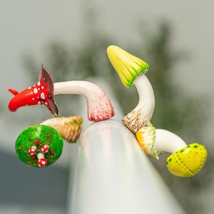Op de afbeelding: Een verzameling grillige paddenstoelbeeldjes in levendige kleuren. De paddenstoelen hebben rode, gele en groene doppen met witte stelen. Eén paddenstoel heeft een rode dop met witte stippen. De beeldjes zijn gerangschikt op een wit oppervlak.