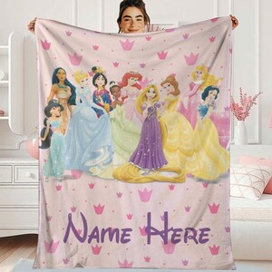 A person is holding a pink blanket with a Disney princess design, featuring various characters such as Rapunzel, Snow White, and other princesses. The blanket has a personalized text that reads "Name Here".