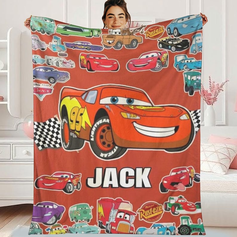 A person is holding a blanket featuring various cars from the Disney Pixar movie Cars, including Lightning McQueen, Rusteze, and other iconic vehicles.