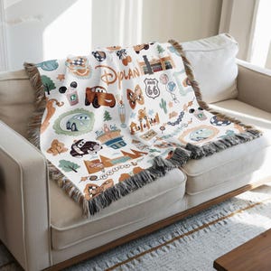A colorful throw blanket with various Disney-themed illustrations, including cars, buildings, and other iconic elements, is draped over a white couch.