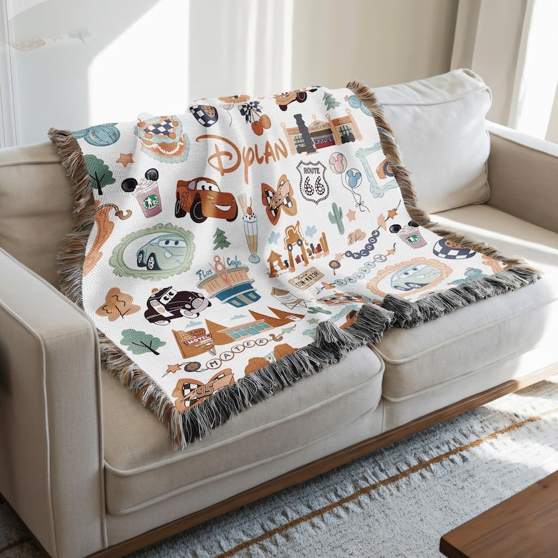 A colorful throw blanket with various Disney-themed illustrations, including cars, buildings, and other iconic elements, is draped over a white couch.
