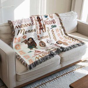 a cozy couch with a colorful, textured blanket or throw on it. The blanket has various cartoon characters and designs, including Moana, a character from the Disney animated film "Moana". The blanket is placed on a white couch, and there is a wooden coffee table in the foreground.