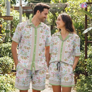 May include: Matching pajama set with a whimsical pattern of green frogs, pink bows, and cherries on a white background. The set includes a short-sleeved button-down shirt and shorts with green trim. The couple is wearing the matching pajama set.