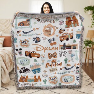 A woman is holding up a colorful, hand-knitted blanket with various Disney-themed designs, including cars, buildings, and other iconic elements from the Cars movie franchise.