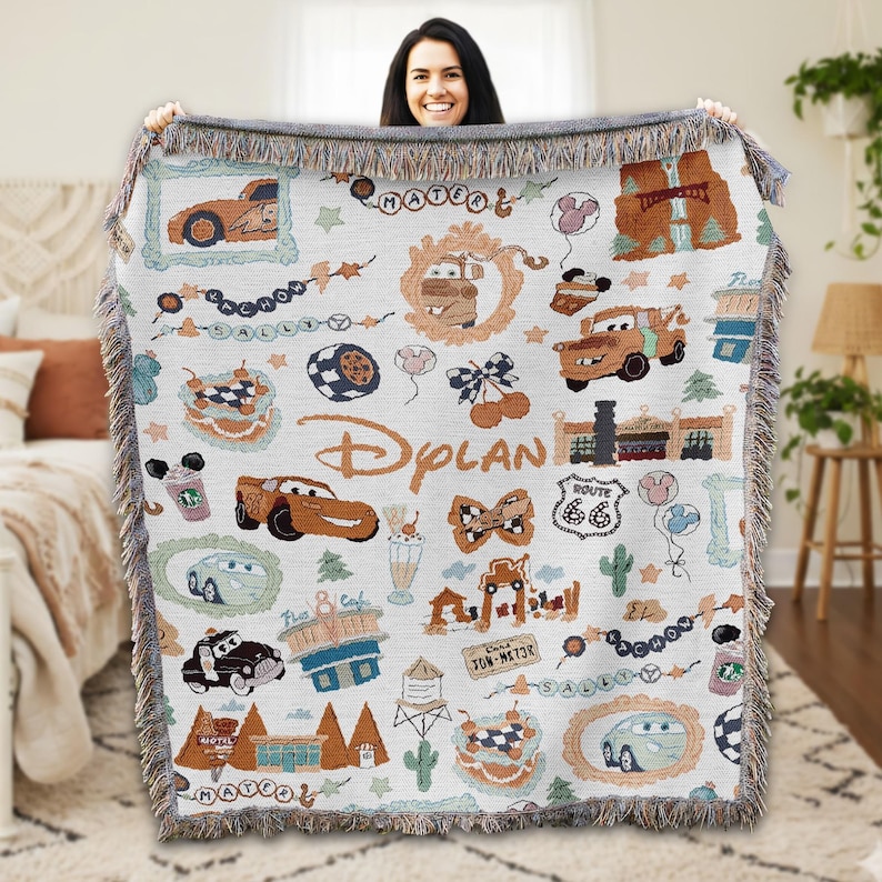 A woman is holding up a colorful, hand-knitted blanket with various Disney-themed designs, including cars, buildings, and other iconic elements from the Cars movie franchise.