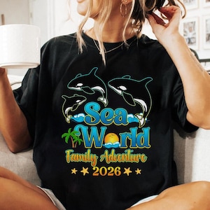 May include: Black t-shirt with a graphic featuring three orcas, the text "Sea World Family Adventure", a palm tree, a sun, and the year 2026. The design is in shades of blue, yellow, and green.