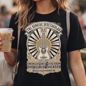 May include: A black short-sleeve t-shirt featuring a graphic of a guitar with a skull, sunburst rays, and text that reads: THE SUNRISE SPECTACULAR, ONE NIGHT ONLY LIVE FROM THE TOWER THEATER, SEIZE YOUR MOMENT. The shirt has a crew neckline.