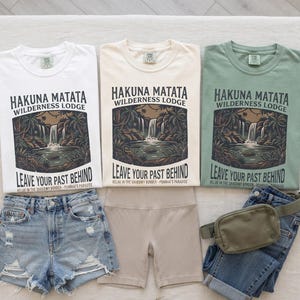 May include: Three t-shirts in white, cream, and sage green featuring a graphic of a waterfall and jungle with the text Hakuna Matata Wilderness Lodge Leave Your Past Behind. Includes distressed denim shorts, beige biker shorts, and a green fanny pack.