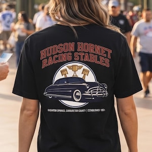Op de afbeelding: Een zwart t-shirt met korte mouwen en een afbeelding op de achterkant van een oldtimer, drie trofeeën en de tekst "HUDSON HORNET RACING STABLES" in het rood, samen met "RADIATOR SPRINGS, CARBURETOR COUNTY | ESTABLISHED 1951" in witte letters.