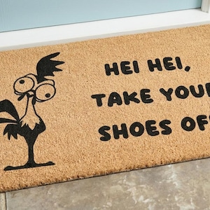 Puede incluir: Un felpudo rectangular de fibra de coco color canela con una ilustraci&oacute;n gr&aacute;fica negra de un gallo de ojos grandes a la izquierda y el texto negro "HEI HEI, TAKE YOUR SHOES OFF" impreso en una fuente negrita y redondeada a la derecha.