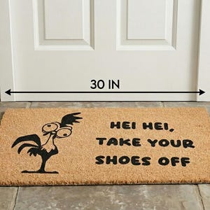 Puede incluir: Un felpudo rectangular de fibra de coco color canela con un gr&aacute;fico negro de un gallo de ojos grandes y el texto "HEI HEI, TAKE YOUR SHOES OFF". La alfombra mide 76,2 cm de ancho y 43,18 cm de profundidad frente a una puerta blanca.