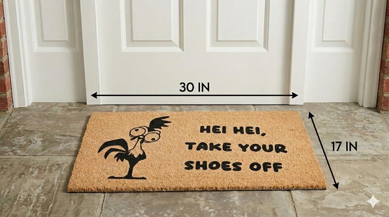 Puede incluir: Un felpudo rectangular de fibra de coco color canela con un gr&aacute;fico negro de un gallo de ojos grandes y el texto "HEI HEI, TAKE YOUR SHOES OFF". La alfombra mide 76,2 cm de ancho y 43,18 cm de profundidad frente a una puerta blanca.