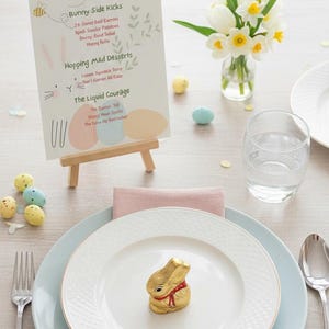 Könnte beinhalten: Oster-Tischdekoration mit einem goldverpackten Schokoladenhasen auf einem weißen Teller. Der Tisch umfasst eine Speisekarte, Blumen und pastellfarbene Teller und Servietten. Die Speisekarte lautet "SOME-BUNNY IS HUNGRY."