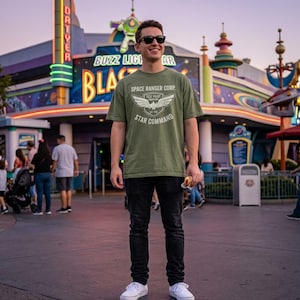 May include: An olive green short-sleeve t-shirt featuring a white graphic print on the chest that reads SPACE RANGER CORP. TEST PILOT STAR COMMAND with a winged emblem. The shirt is paired with black denim jeans and white canvas sneakers.