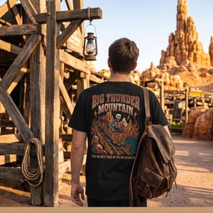 May include: A black t-shirt featuring a graphic print on the back with the text Big Thunder Mountain and The Wildest Ride In The Wilderness, depicting a skeleton in a mine cart, flames, and mountains, paired with a brown leather backpack.