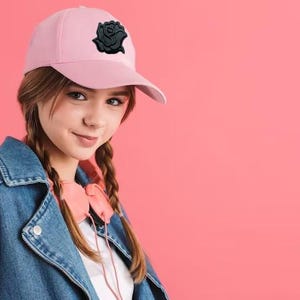 Puede incluir: Gorra de b&eacute;isbol rosa claro con un emblema de rosa negra. La persona lleva una chaqueta vaquera y auriculares rosas. El fondo es de color rosa liso.