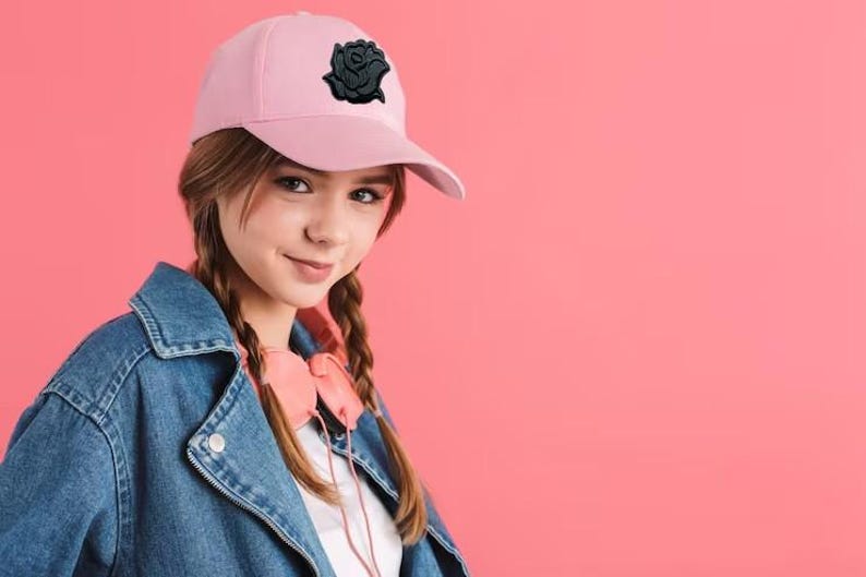 Puede incluir: Gorra de b&eacute;isbol rosa claro con un emblema de rosa negra. La persona lleva una chaqueta vaquera y auriculares rosas. El fondo es de color rosa liso.