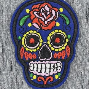 Peut inclure: Un écusson brodé représentant un crâne de style calavera bleu. Le crâne est orné d'une grande rose rouge sur le front, d'orbites en forme de fleurs jaunes avec des centres noirs, d'accents orange sur les joues et de dents blanches. Le motif est souligné par un fil noir sur une base en tissu bleu.