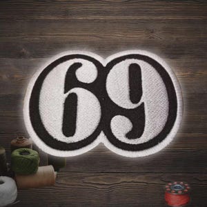 May include: Embroidered patch featuring the number "69" in white, outlined in black, and set against a black background. The patch has a white border and is likely designed to be sewn or ironed onto fabric. The design is centered on a wooden surface.