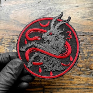 May include: A circular embroidered patch featuring a black goat with red eyes and horns, surrounded by a red snake and three red teardrop shapes. The design includes a red crescent moon and the number three in red on a black background.