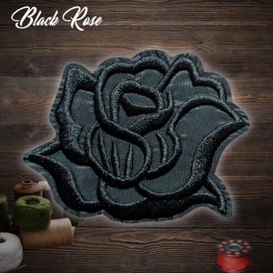 Puede incluir: Parche de rosa negra bordada con el texto "Black Rose" en cursiva. El parche es negro con costuras detalladas, sobre un fondo de madera. La rosa es el punto focal.