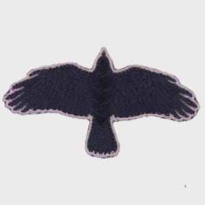 May include: An embroidered patch in the shape of a bird in flight, featuring a dark navy blue body and wings with a light lavender or pale purple stitched border outlining the entire silhouette of the bird with its wings fully extended.
