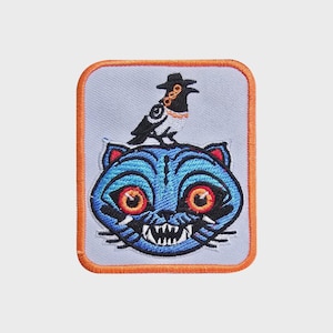 May include: Embroidered patch with a blue cat and a bird wearing a hat. The cat has red eyes and sharp teeth. The patch has an orange border and a light blue background. The design is a quirky illustration.