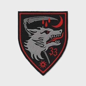 Puede incluir: Un parche bordado en forma de escudo que presenta una cabeza de lobo gris de perfil con una lengua roja, una guadaña gris encima, tres gotas de sangre rojas, una luna creciente roja, el número 33 rojo y una estrella roja sobre fondo negro.