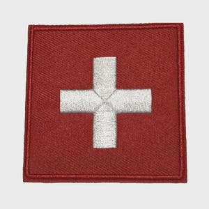 May include: Embroidered patch featuring the Swiss flag. The patch has a red background with a white cross in the center. The cross is centered and has arms of equal length. The patch is square with a stitched border.