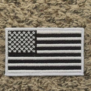May include: A rectangular patch featuring a stylized American flag design. The flag has black and white stripes and a field of white flower-shaped stars. The patch is bordered with white stitching.