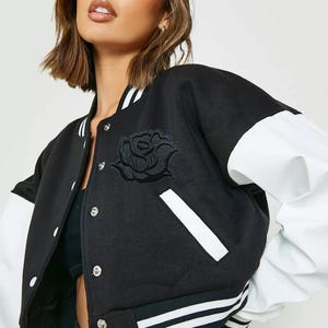 Puede incluir: Chaqueta universitaria negra y blanca con un emblema de rosa. La chaqueta tiene un cuerpo negro, mangas blancas y rayas blancas en los pu&ntilde;os y el dobladillo. Tiene una parte delantera abotonada y un parche de rosa en el pecho.
