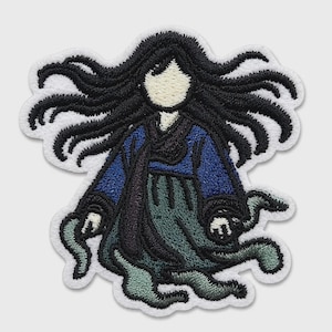 May include: An embroidered patch featuring a figure with long, flowing black hair, wearing a dark blue top and a teal skirt. The figure has a pale, featureless face and appears to be floating with wispy, trailing edges at the bottom of the garment.