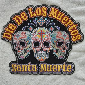 Peut inclure: Un écusson brodé noir représentant trois crânes en sucre avec des motifs floraux et des croix colorés. Le texte "Dia De Los Muertos" est arqué en jaune au-dessus des crânes, et "Santa Muerte" est écrit en jaune en dessous sur fond noir.