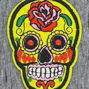 Peut inclure: Un écusson brodé représentant un crâne en sucre jaune vif. Le crâne est orné d'une grande rose rouge sur le front, d'orbites en forme de fleurs jaunes avec des centres orange, d'un nez noir et d'une mâchoire rose avec des dents blanches.