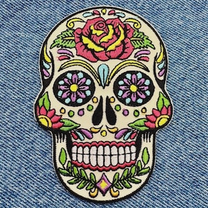 Peut inclure: Un écusson brodé représentant un crâne en sucre de couleur crème avec des détails floraux vibrants. Le motif comprend une grande rose rose et jaune sur le front, des orbites en forme de fleurs violettes et des feuilles vertes, le tout bordé de noir.