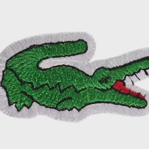 Op de afbeelding: Geborduurde patch met een groen krokodil met een rode mond, omlijnd in zwart en met een witte rand. De patch is een herkenbaar logo, vaak te vinden op kleding en accessoires. Het ontwerp is een gedetailleerde, getextureerde weergave van het dier.