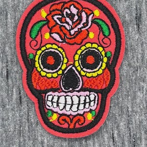 May include: An embroidered patch featuring a red sugar skull design with a large pink and orange rose on the forehead, yellow eye sockets, a black nose, and white teeth. The skull is outlined in black thread against a red felt background.