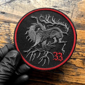 May include: A circular embroidered patch featuring a black background with a red border. The design depicts a grey mythical creature with elongated limbs and a long tail, positioned above the red embroidered number 33 in a gothic font style.