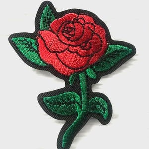 May include: An embroidered patch featuring a vibrant red rose with detailed green leaves and a green stem. The design is set against a black background, with the entire shape of the flower and foliage outlined in a black border for clear definition.
