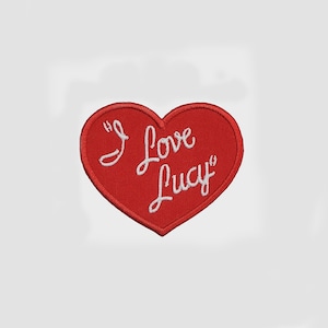 May include: A heart-shaped embroidered patch with a red fabric base and a stitched red border. The text "I Love Lucy" is embroidered in white cursive script across the center of the heart, featuring a clean, textured finish throughout.