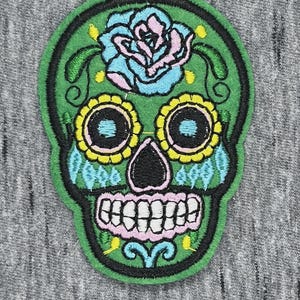 Peut inclure: Un écusson brodé représentant un crâne en sucre vert. Le crâne présente une rose bleu clair sur le front, des orbites jaunes avec des centres bleus, un nez noir et une mâchoire rose avec des dents blanches, le tout souligné par un fil noir épais.