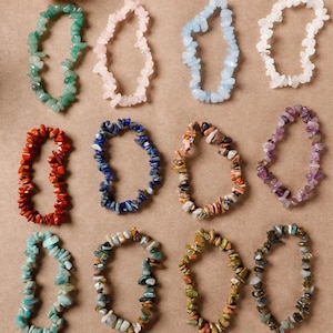 May include: A collection of twelve gemstone bracelets in various colors, including green, pink, blue, white, red, and purple. Each bracelet is made of small, irregularly shaped stones strung together on an elastic band.