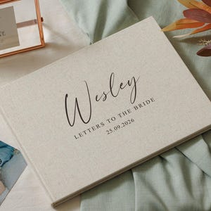 Puede incluir: Un libro rectangular cubierto de tela color crema con el texto impreso en negro "Wesley LETTERS TO THE BRIDE 25.09.2026" en la portada. El libro está colocado junto a un letrero con marco de cobre, una fotografía y un arreglo floral.