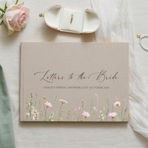 Puede incluir: Un libro de tapa dura beige titulado Letters to the Bride con el texto Grace's Bridal Shower 12th October 2026 impreso en negro. La parte inferior presenta una ilustración en acuarela de flores silvestres en tonos rosa, amarillo y verde.