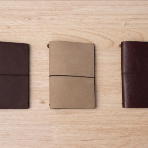 May include: Three leather journals in shades of brown and tan are arranged on a light wood surface. Each journal is closed and secured with an elastic band. The journals are rectangular and have a simple, minimalist design.
